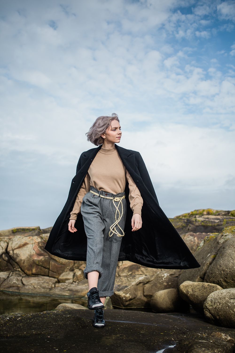 A person in a black coat and beige sweater walks dramatically across rocky coastal terrain against a blue cloudy sky.