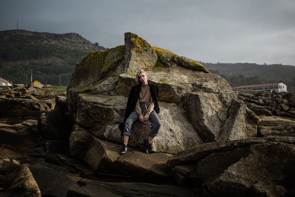A figure sits on large coastal boulders against a moody gray sky with mountains in the background.