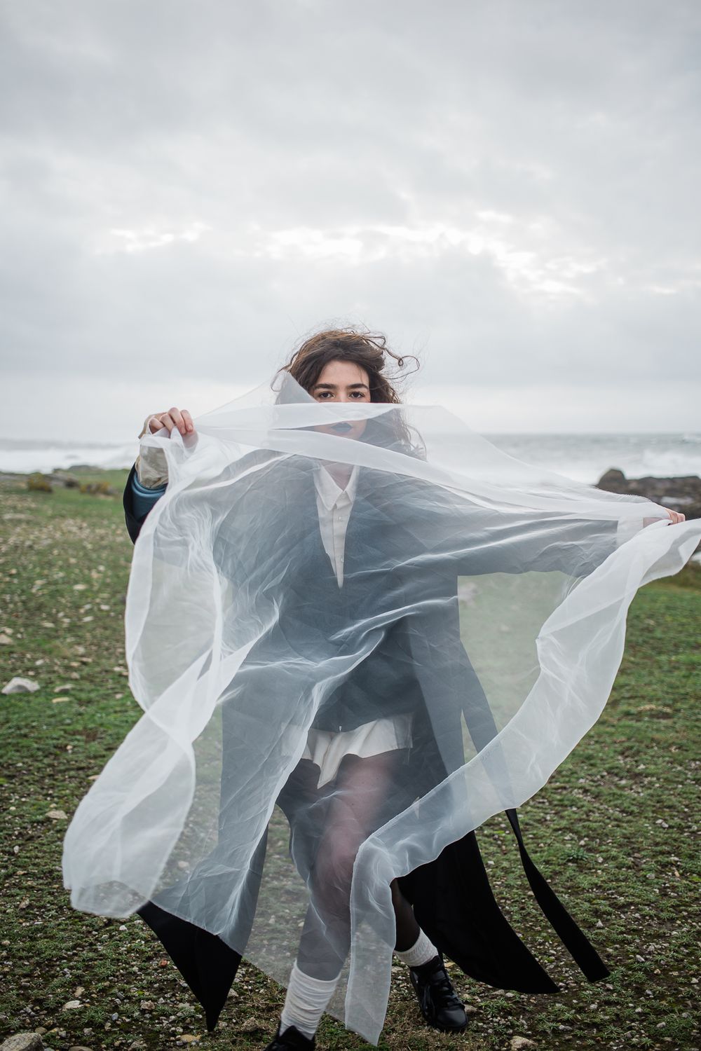 Figure draped in flowing sheer white fabric poses dramatically against cloudy coastal landscape on grassy terrain.