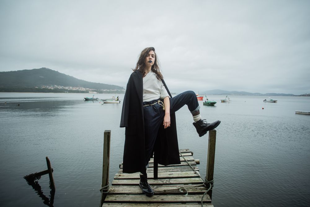 A person in dark coat stands confidently on a wooden dock overlooking a misty harbor with boats in the background.