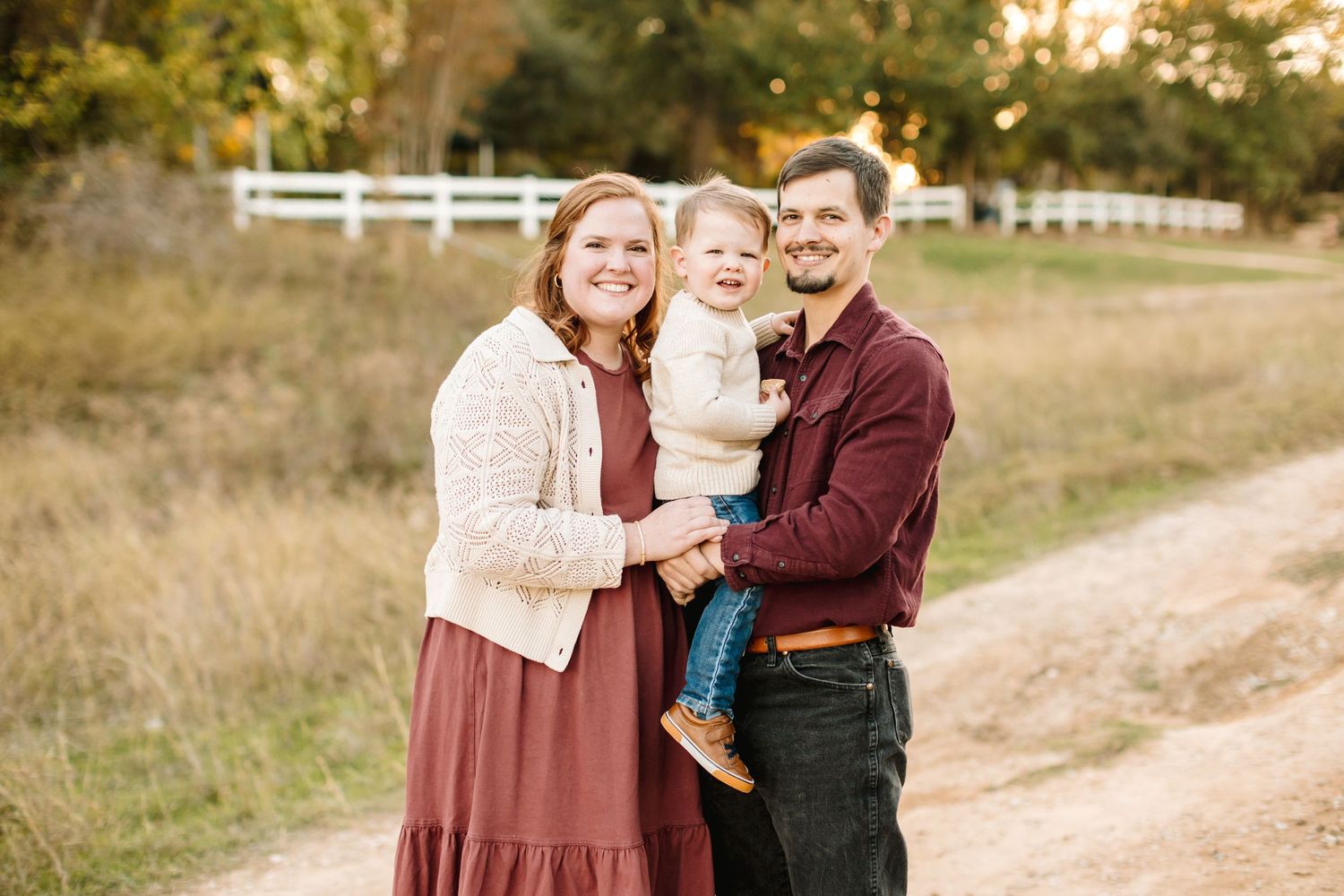 Family portrait session on dirt path with white fence and autumn field in background.