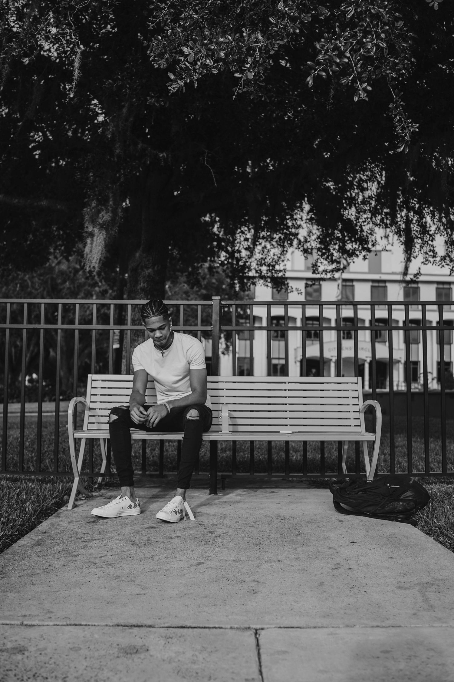 Black and white photo of someone sitting alone on a white bench beneath overhanging trees in a park setting.