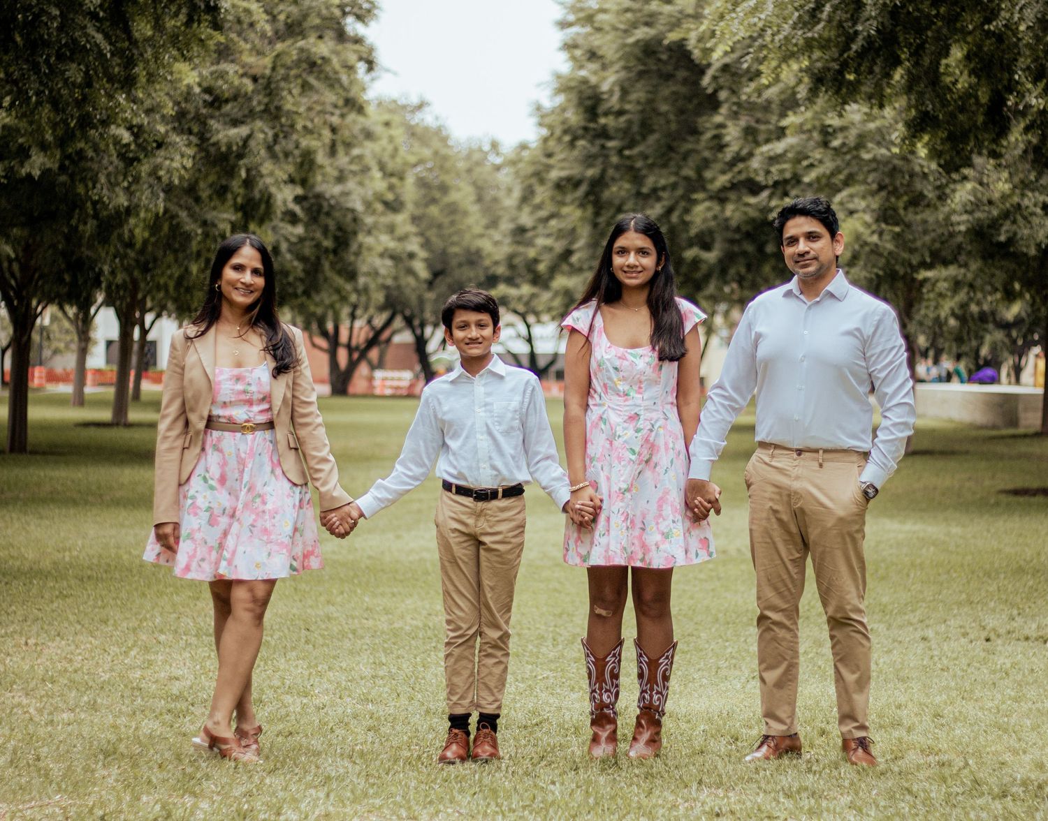 A family of four stands together holding hands in a park setting wearing coordinated spring outfits.