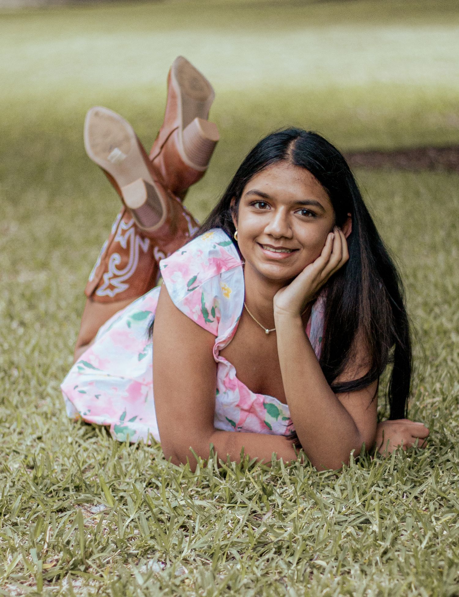 Someone relaxes on grass wearing a floral dress and brown cowboy boots in a dreamy summer portrait.