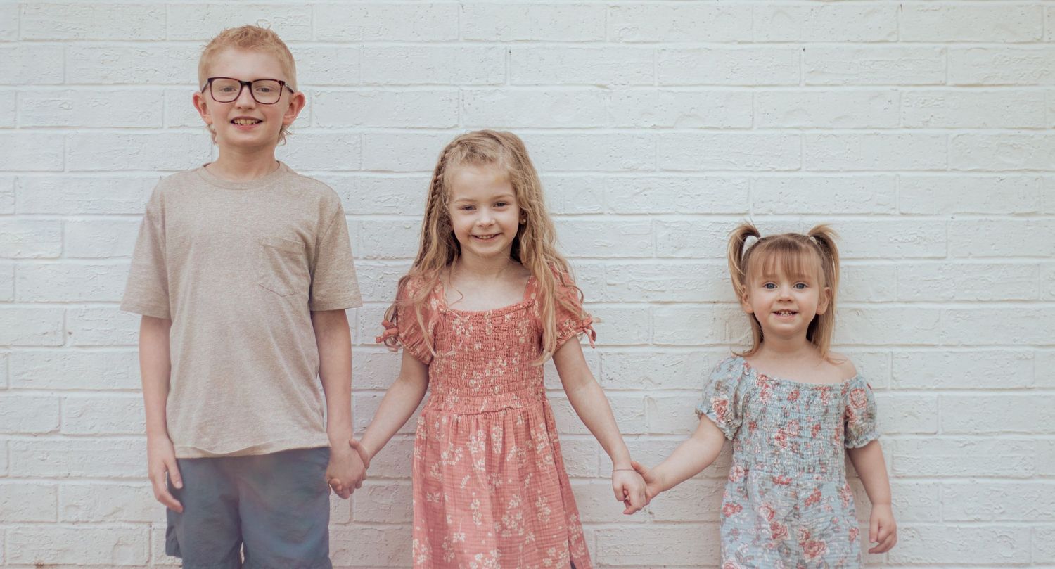 Three young children stand holding hands against a white brick wall, wearing casual summer outfits.