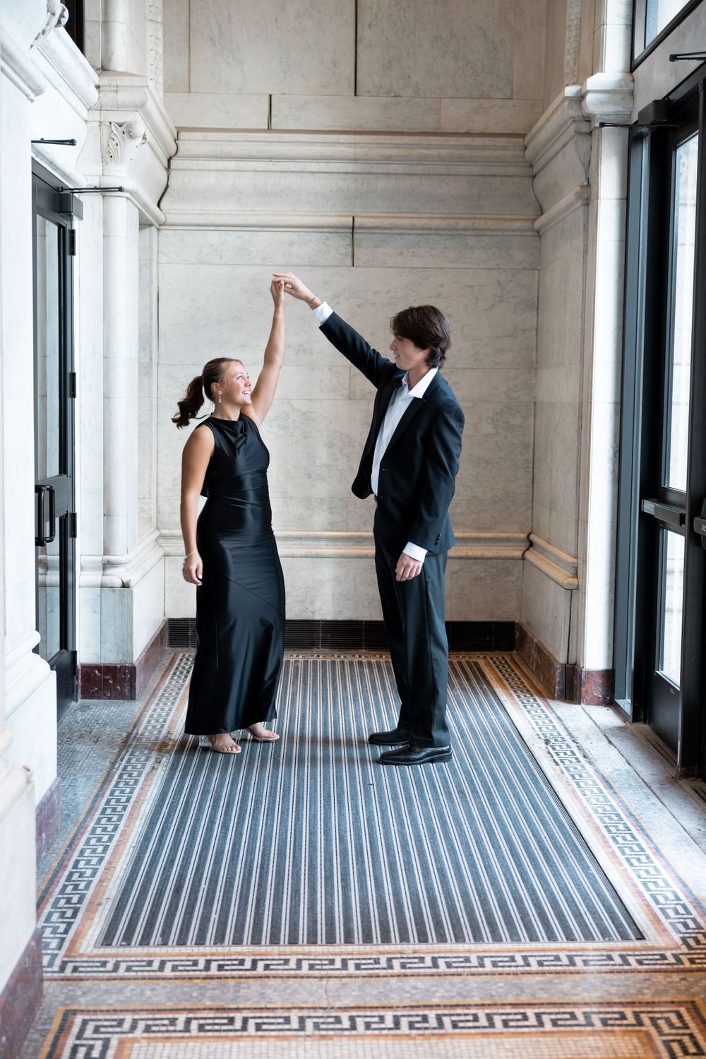 Couple dancing with raised arms in ornate marble hallway with chandeliers.