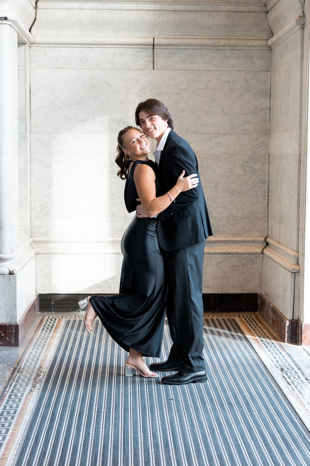Couple embracing in formal attire on a striped rug in an elegant interior with white walls and ornate molding.