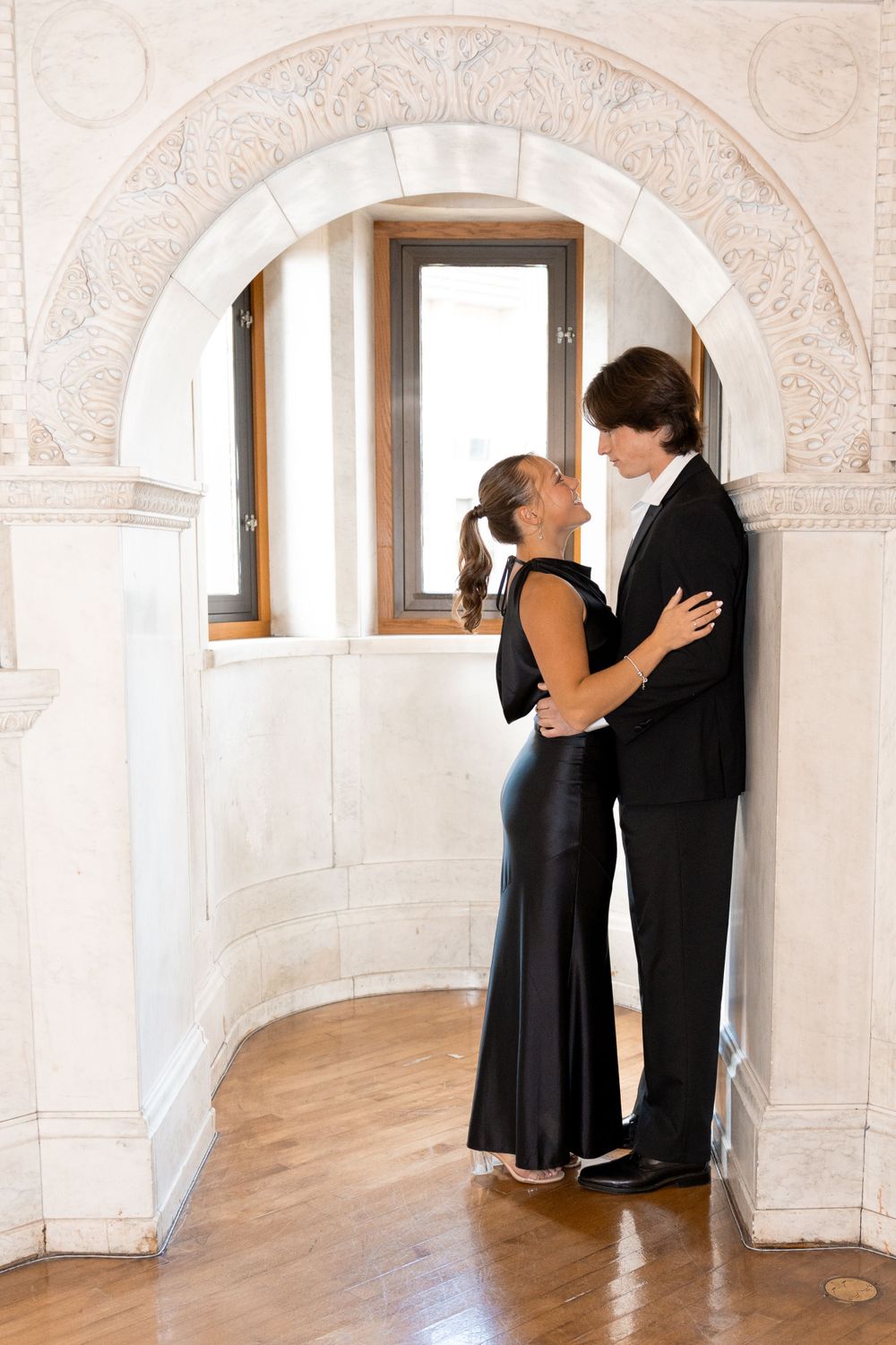 Couple in formal wear standing close together under a decorative archway with windows in the background.