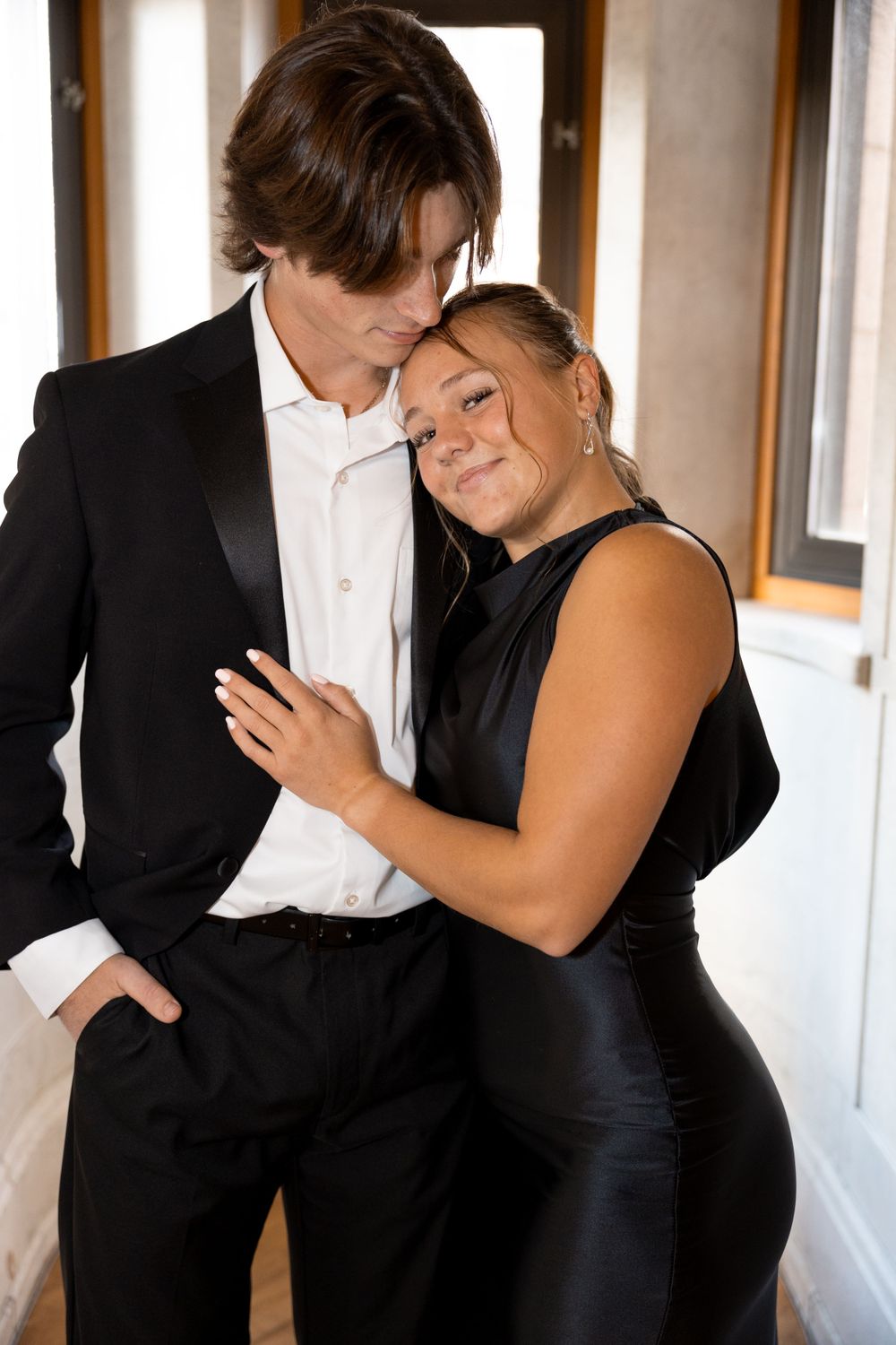 Couple in formal wear hugging in a hallway with windows and white walls.