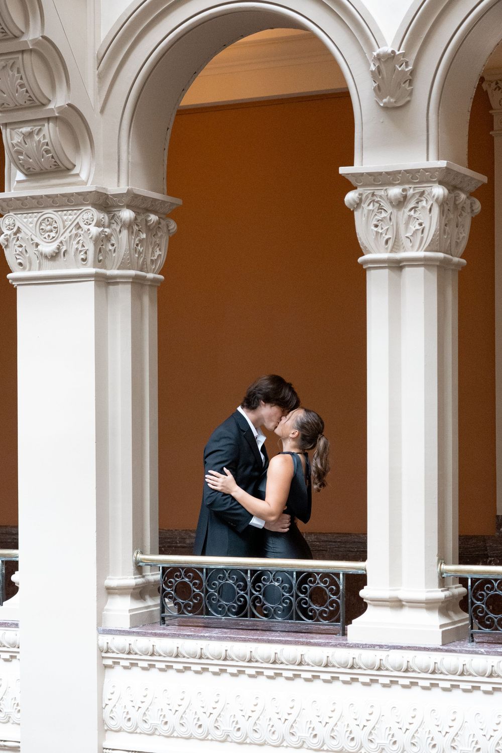 Couple embracing on a balcony framed by ornate white arches and columns.