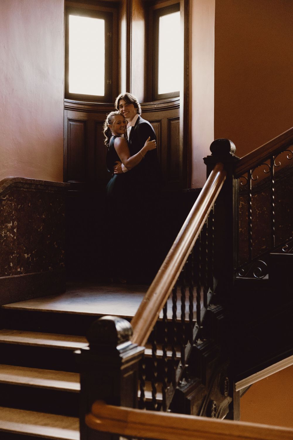 Couple embracing in a window alcove at the top of a staircase with ornate wooden banisters.