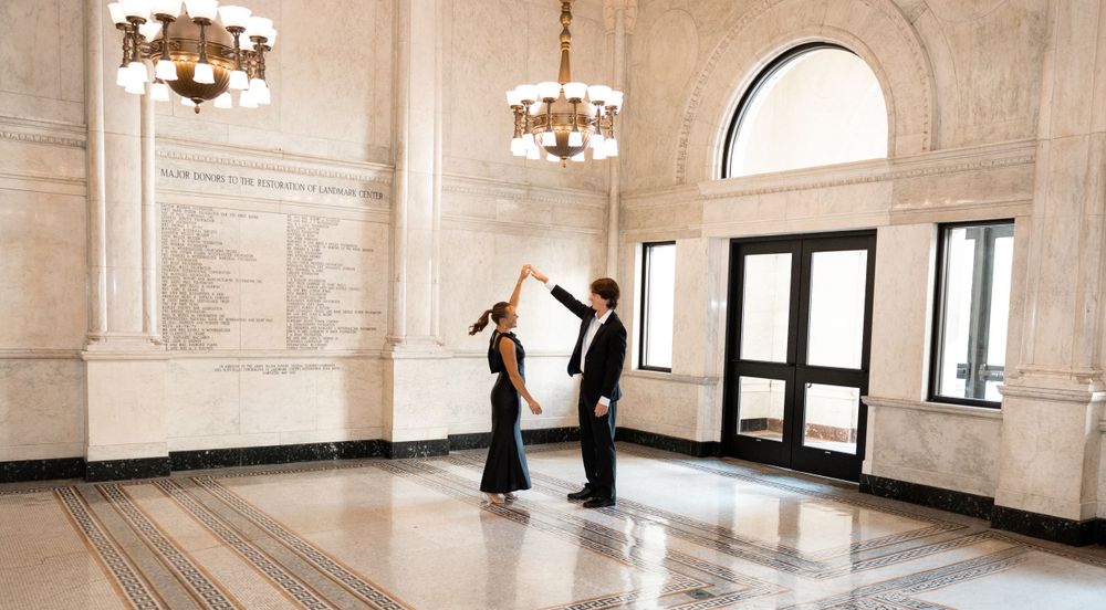 Couple dancing in formal attire in elegant marble hall with chandeliers.
