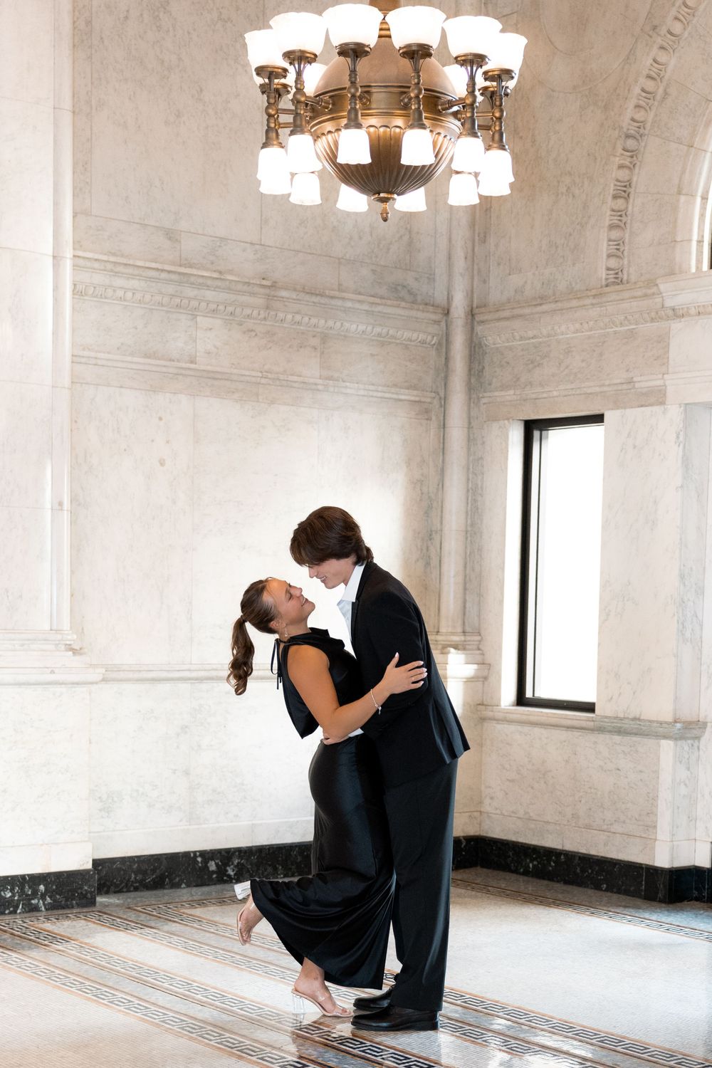 A couple in formal attire dances intimately in an elegant marble interior with a grand chandelier overhead.