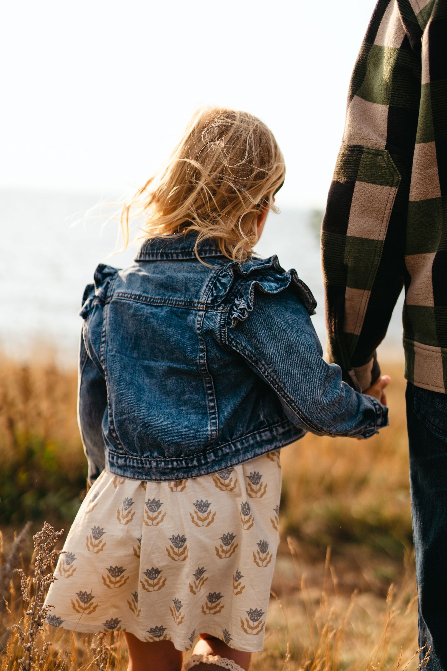 Young child in denim jacket and patterned dress holds hand while walking through tall grass at golden hour.