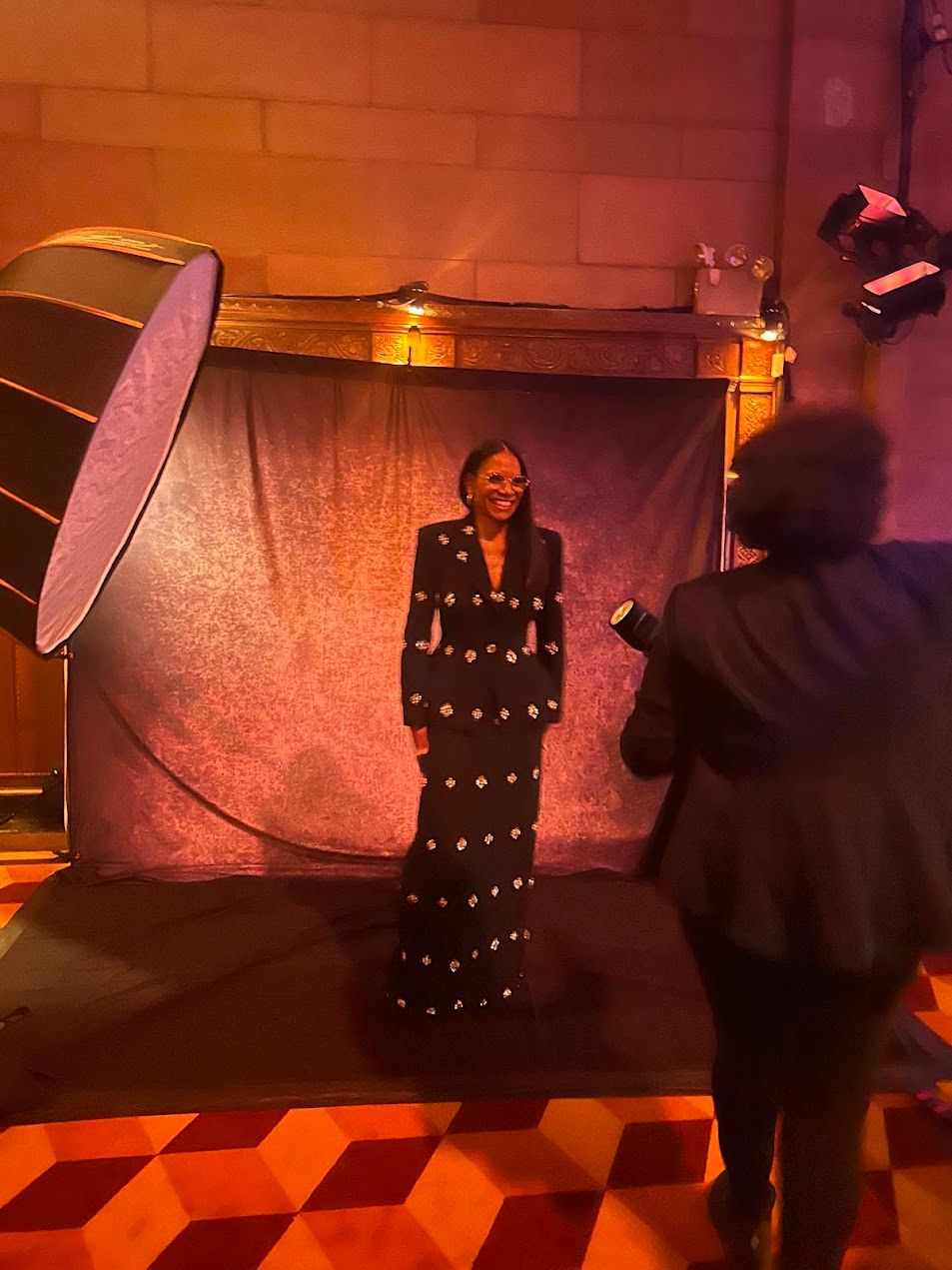 Woman in black embellished gown poses for professional photographer at elegant event with studio lighting.