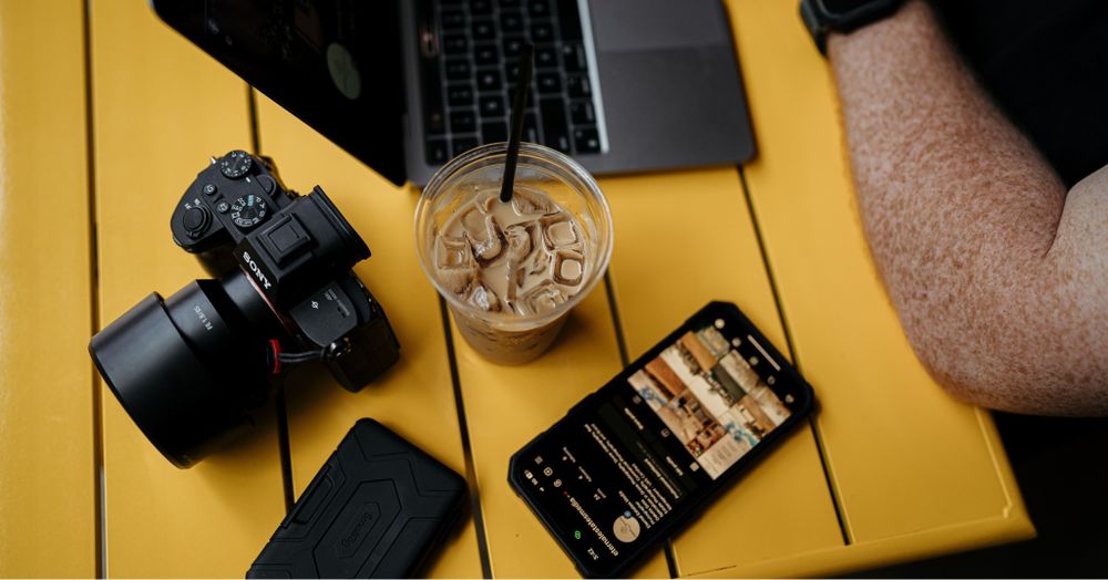 Work setup with camera, iced coffee, laptop and phone on yellow surface.