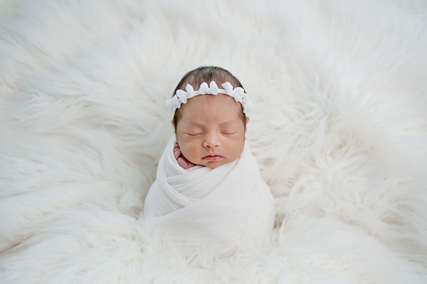 Peaceful newborn swaddled in white wrap with delicate lace headband on fluffy white fur backdrop.