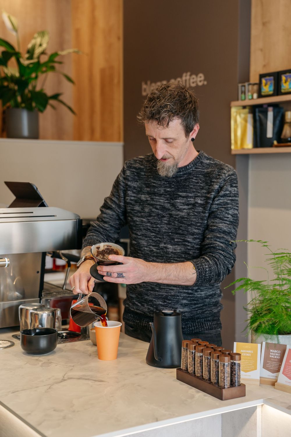 Barista pouring coffee at a modern cafe counter with espresso machine and coffee products displayed.