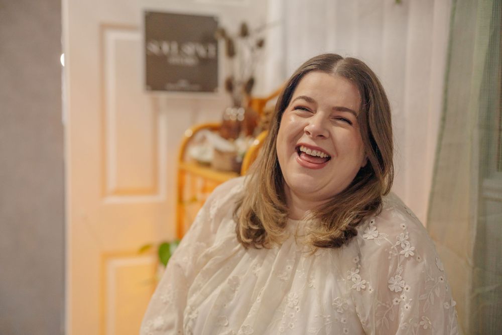 A joyful person with long brown hair laughs while sitting in a warmly lit room with a window in the background.