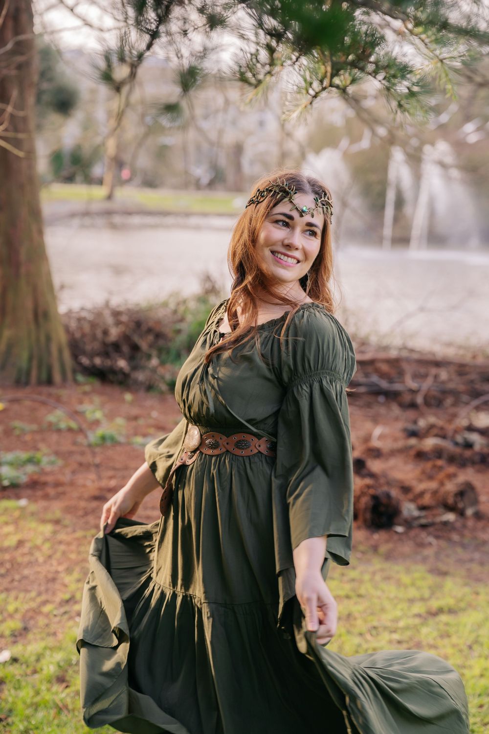 A woman in a flowing dark green dress twirls gracefully in an outdoor woodland setting during autumn.