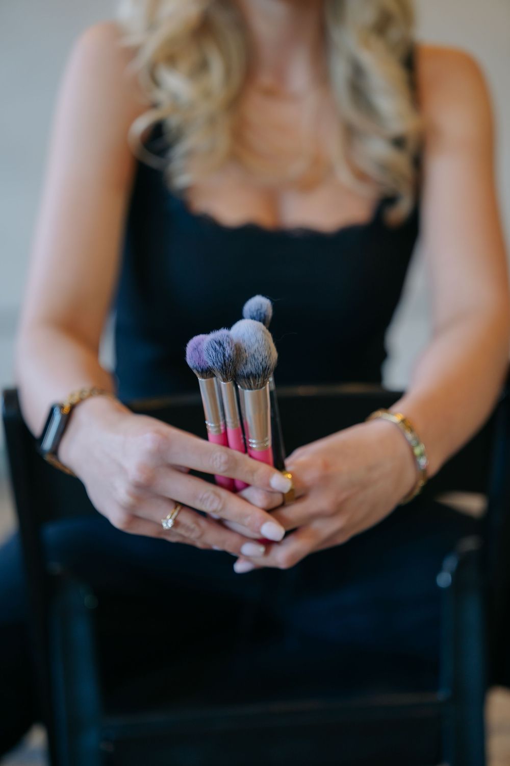 Woman in black dress holding multiple makeup brushes with pink handles, wearing gold bracelets and rings.