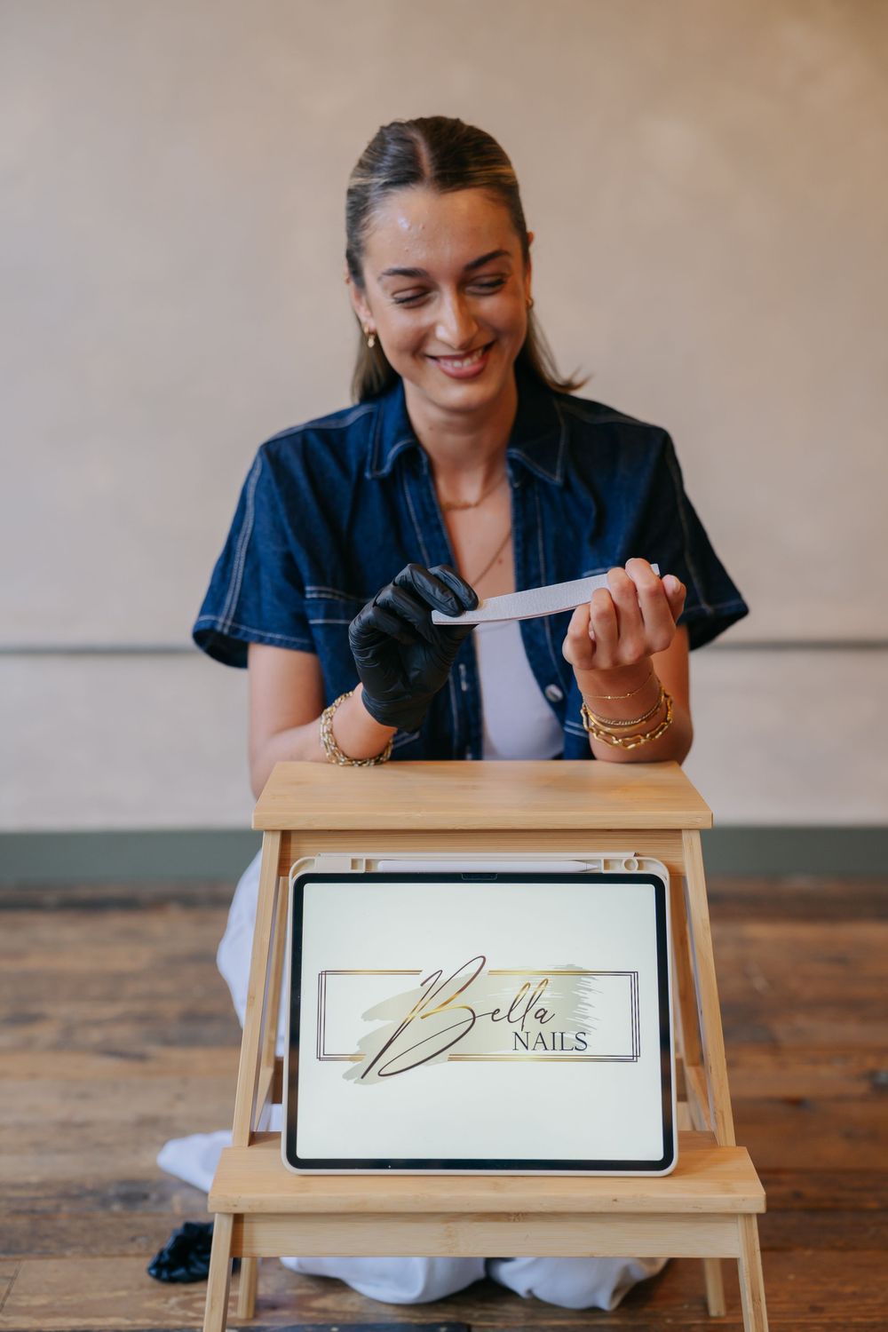 Smiling woman in denim jacket filing nails beside a wooden stool displaying a Bella Nails branded tablet sign.