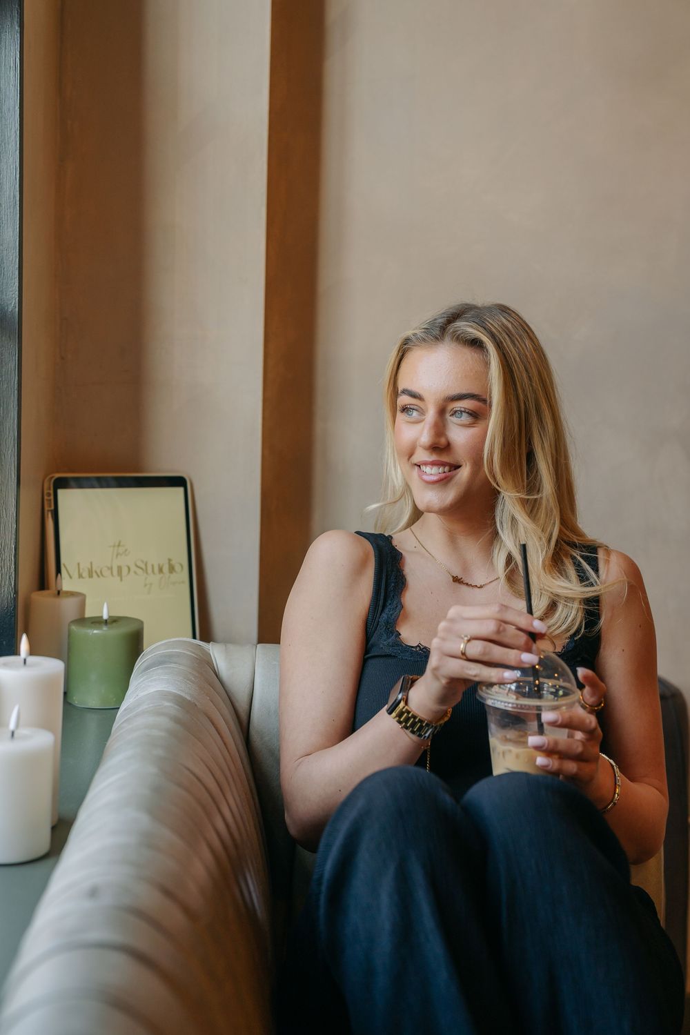 Smiling blonde woman sitting on a sofa holding an iced coffee drink next to candles and a tablet.