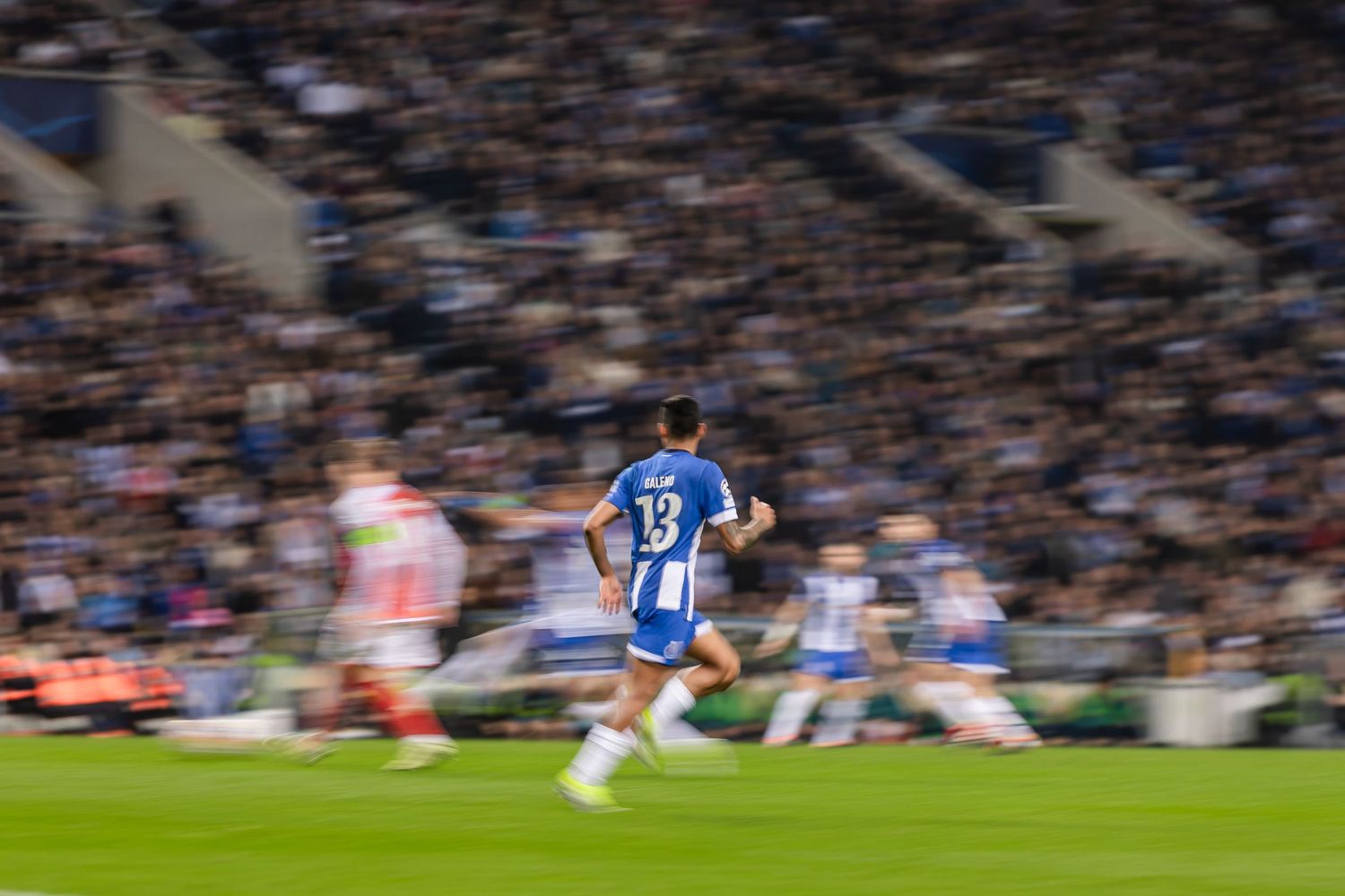 Motion blur photograph of soccer players during a match with a packed stadium crowd in the background.
