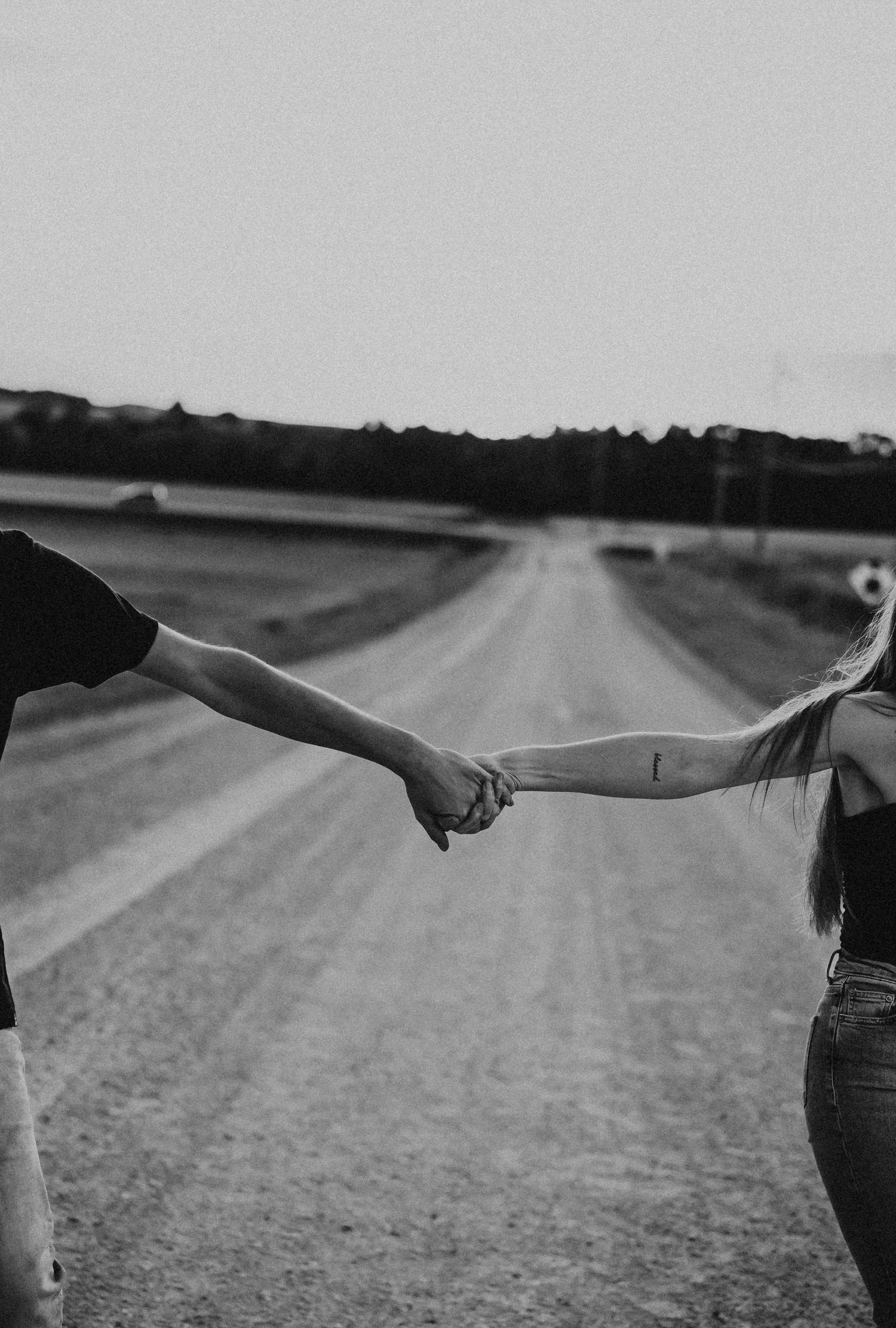 Black and white photo of two people holding hands on a rural dirt road stretching into the distance.