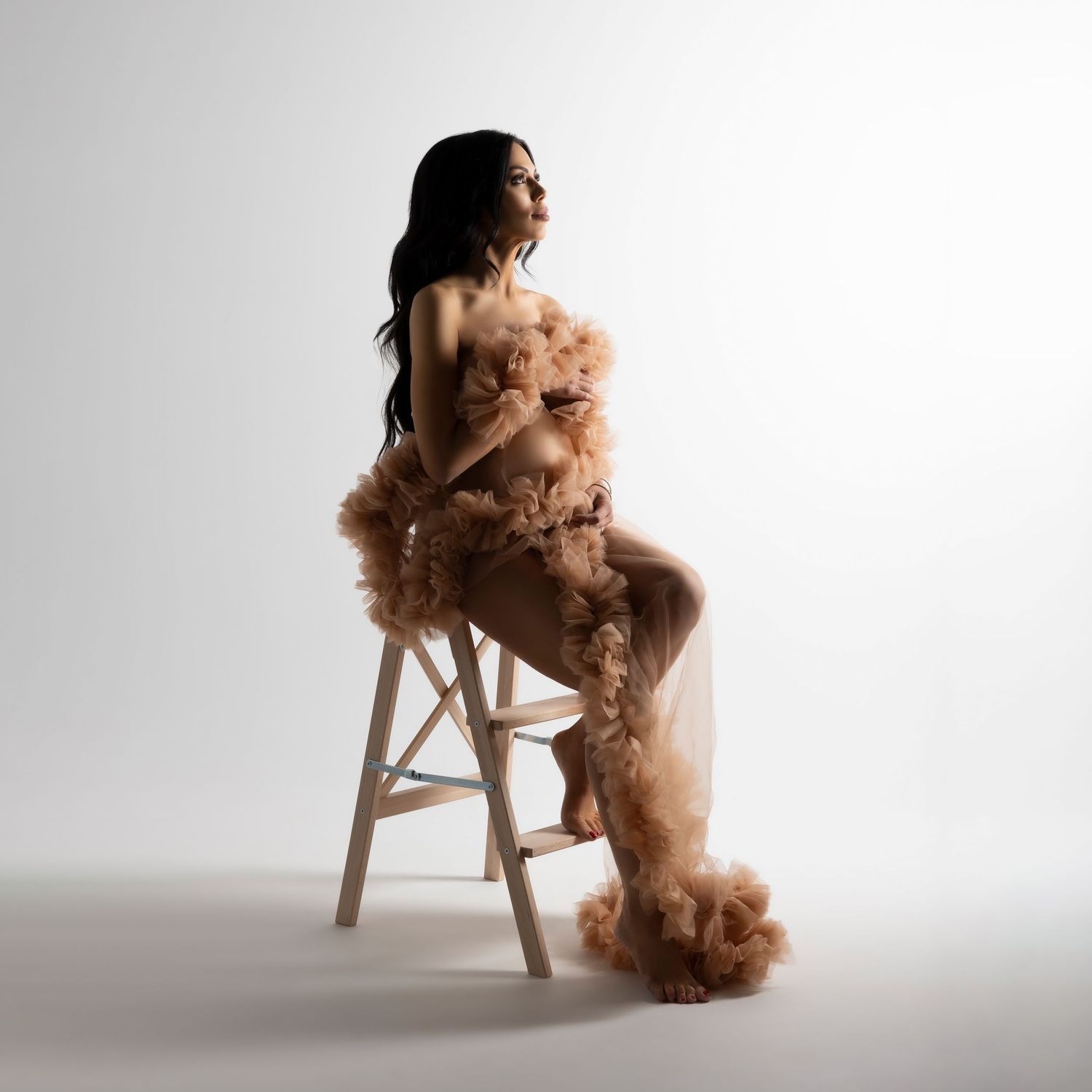 Figure seated on wooden stool draped in flowing peach tulle fabric against white studio background.