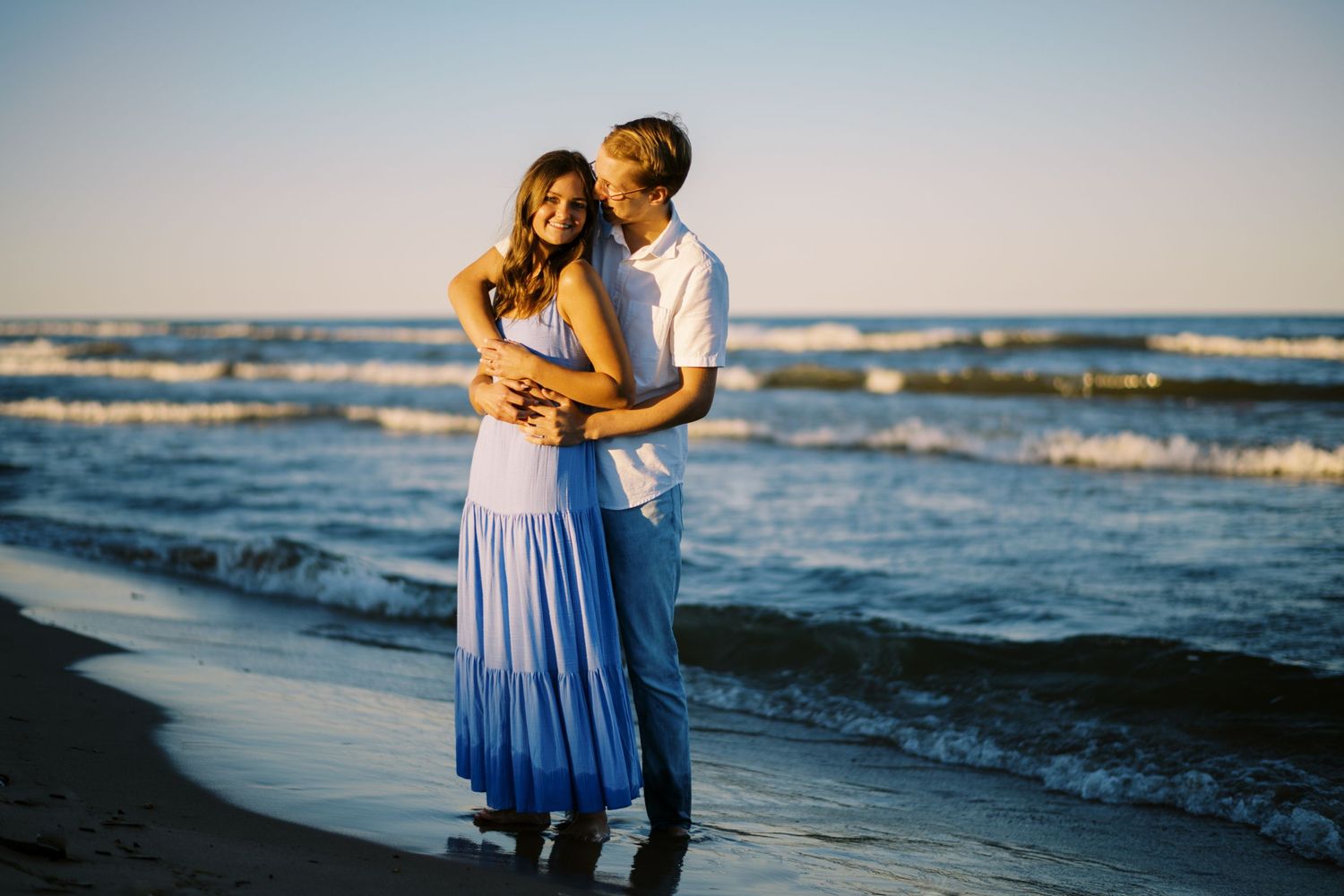 A couple embraces on a beach at sunset while waves crash against the shore in romantic golden light.