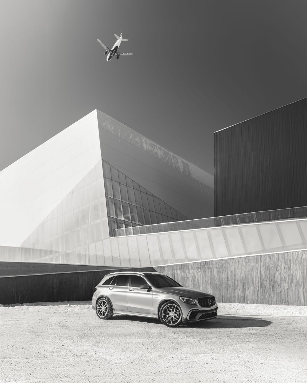 A silver Mercedes SUV parked in front of modern angular architecture with a small airplane flying overhead in black and white.