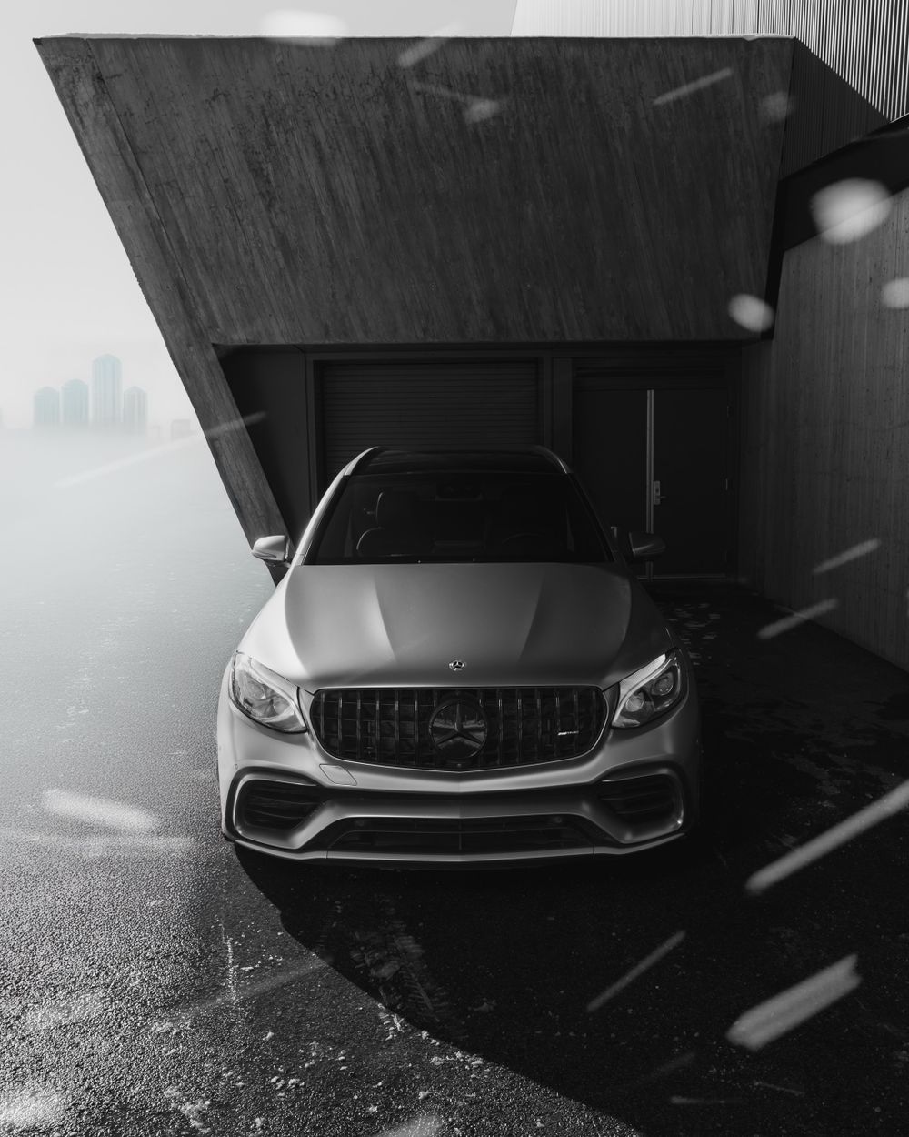 Mercedes-Benz AMG GLC63 in black with glowing headlights parked under dark overhang on rainy day with foggy skyline.