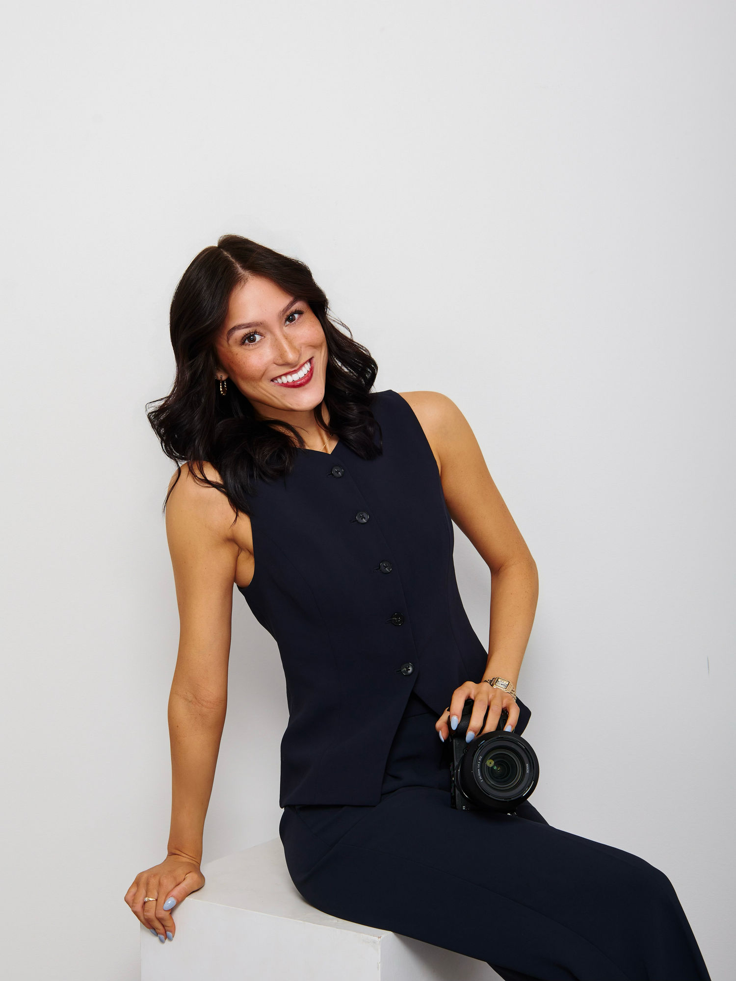 Professional portrait of a woman wearing a black blazer and top with delicate necklace, smiling warmly against neutral background.