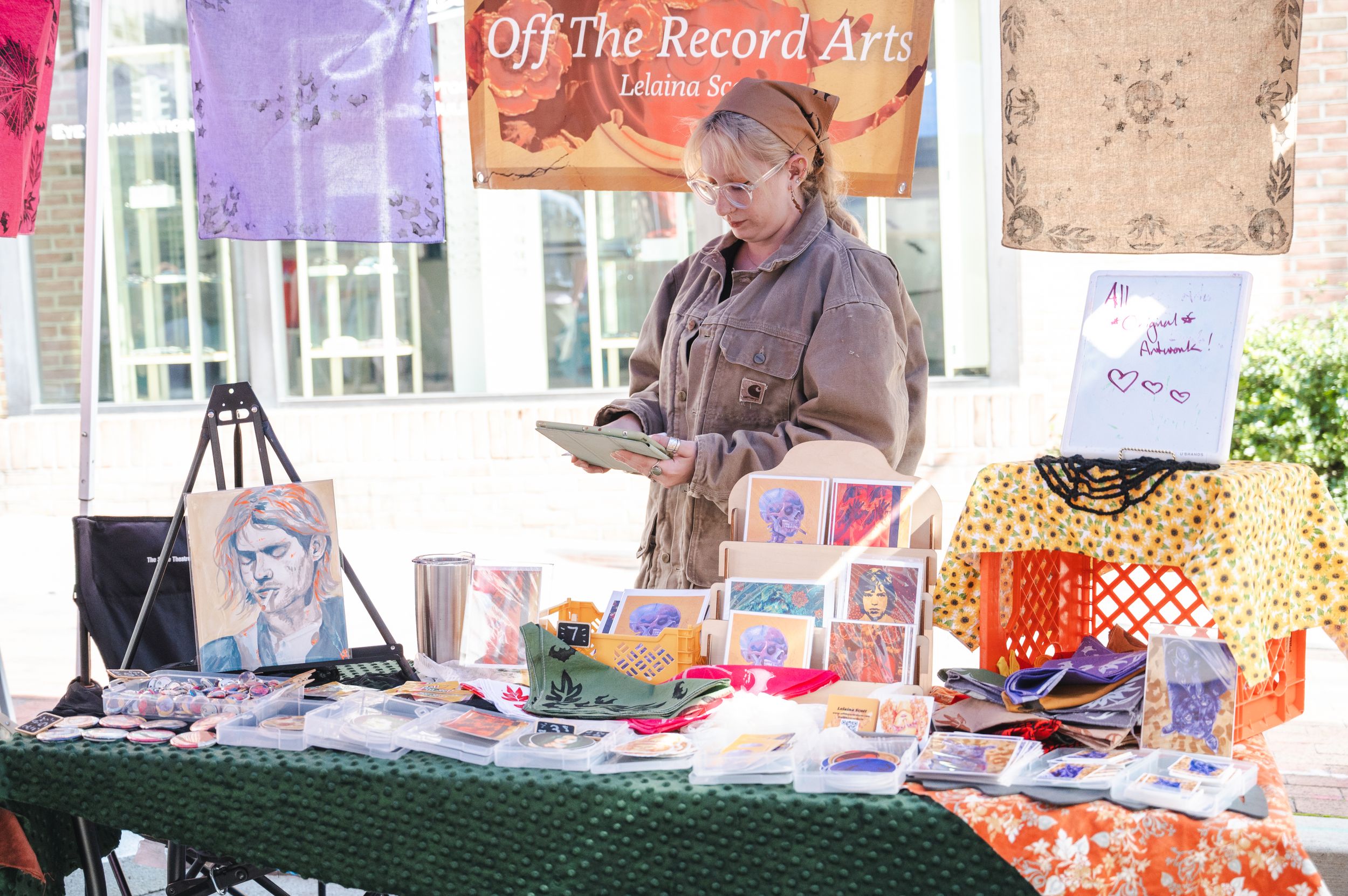 Vendor displaying artwork and merchandise at outdoor market booth with purple banner.