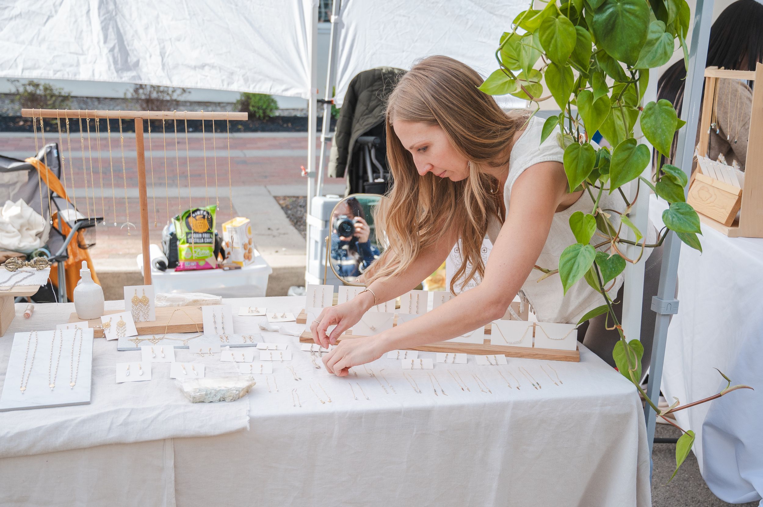 Craft vendor arranging items on white tablecloth display at outdoor market.