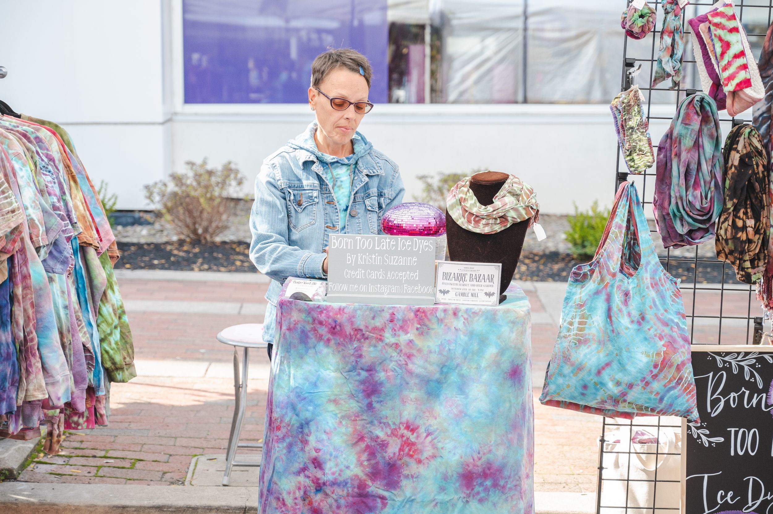 Vendor displaying tie-dye clothing and accessories at outdoor market stall.