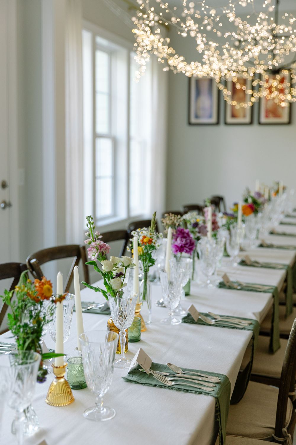 Long wedding reception table with string lights and colorful floral arrangements.