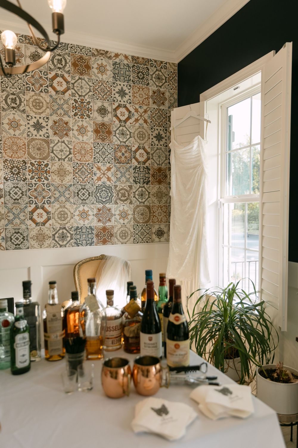 Bar setup against decorative tile wall with copper mugs and bottles near window with white curtain.