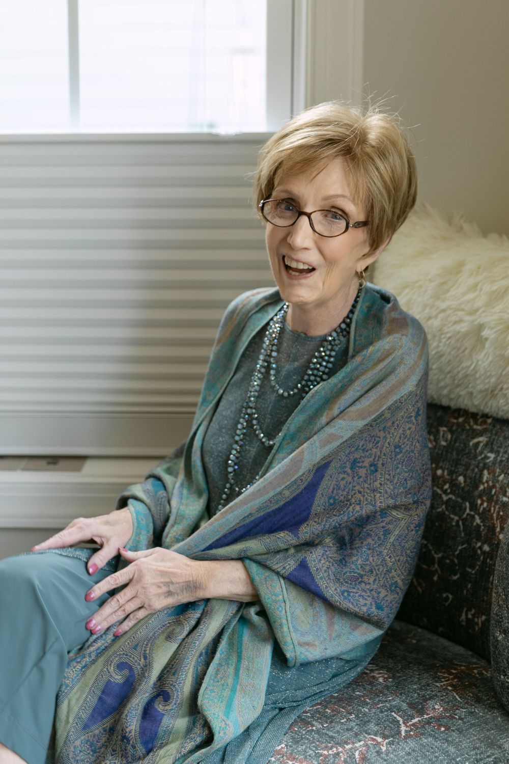 A seated figure wearing glasses and a turquoise shawl smiles warmly while relaxing on a couch by a window.