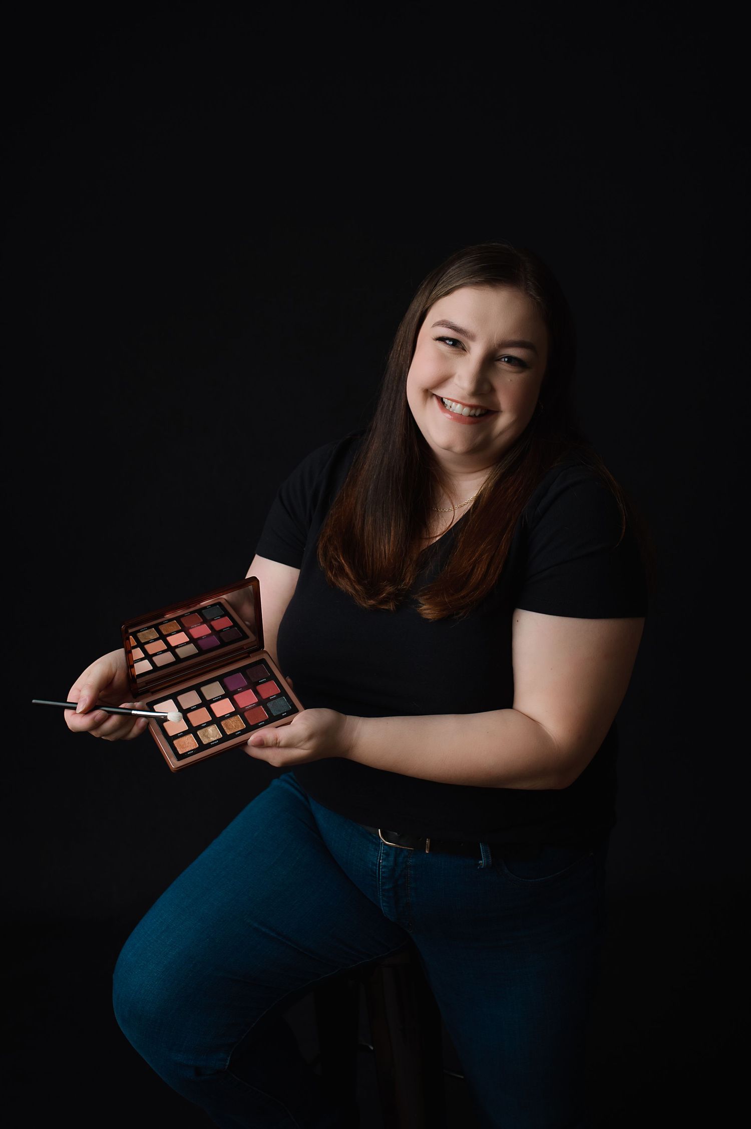 Young woman in black shirt holds a makeup eyeshadow palette against dark background with joyful expression.