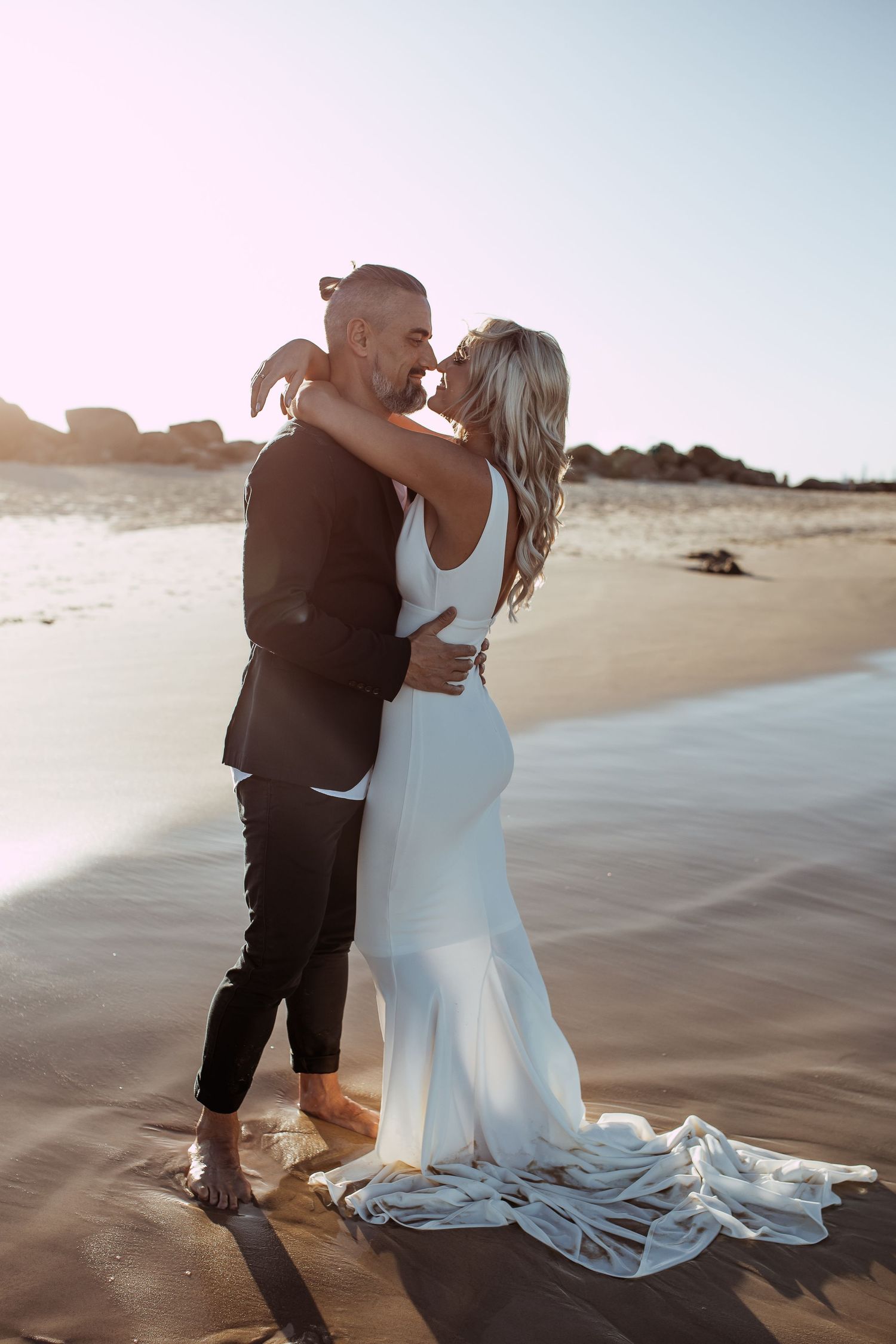 A romantic couple embraces on a sunlit beach, the bride wearing a flowing white gown with a long train in the sand.