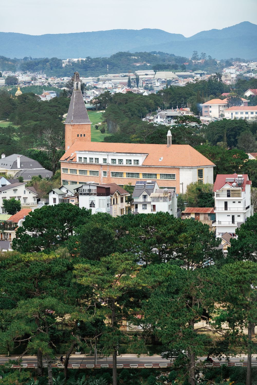 Cityscape view of a hillside town with a prominent church spire and orange-tiled roofs among lush greenery.