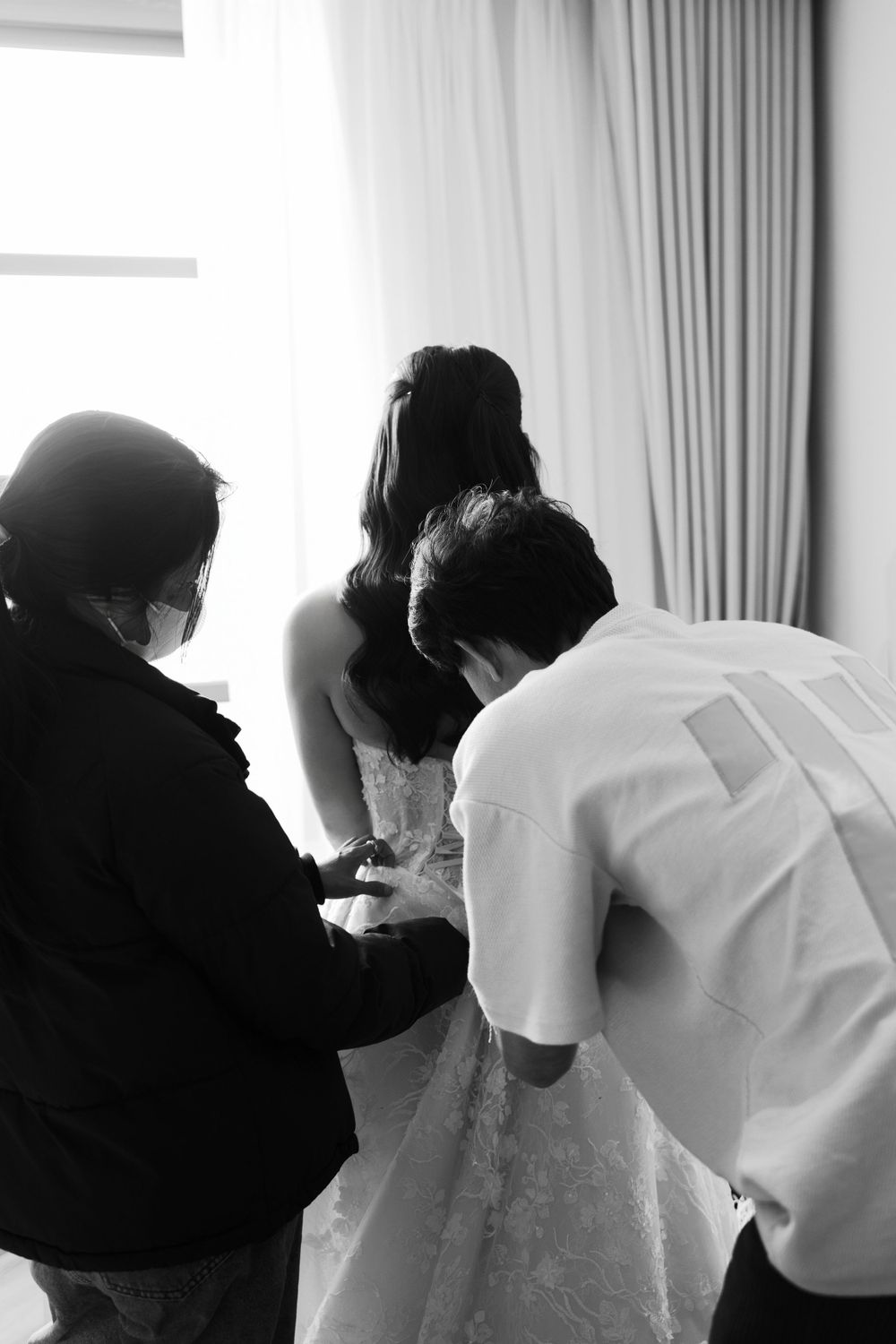 Black and white photo showing final wedding dress preparations with helpers making adjustments.