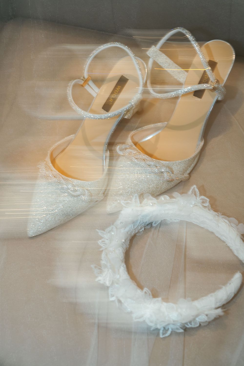 Delicate white lace bridal shoes and floral headpiece arranged on light fabric.