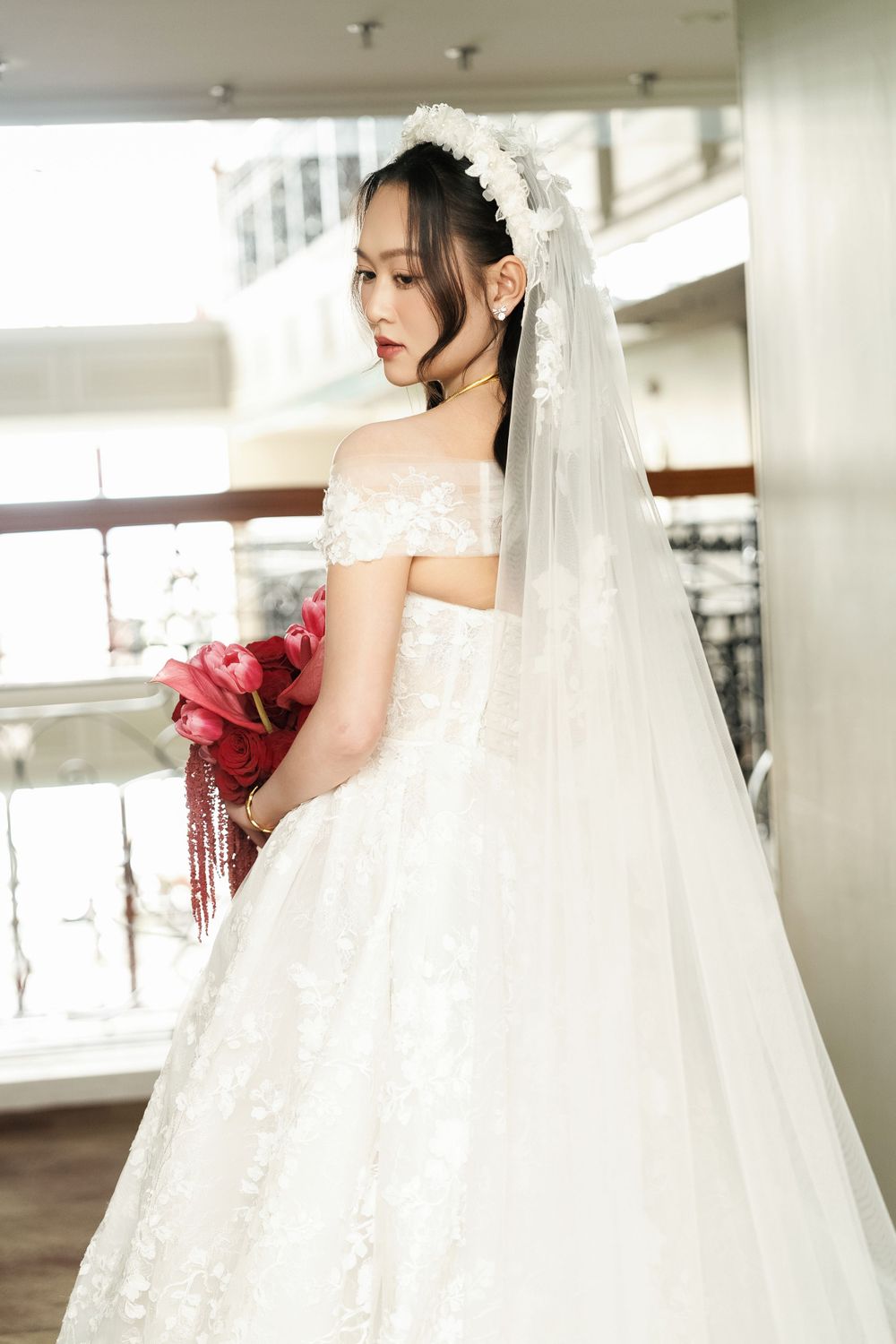 Bride in an off-shoulder lace wedding gown holding red bouquet against white interior.