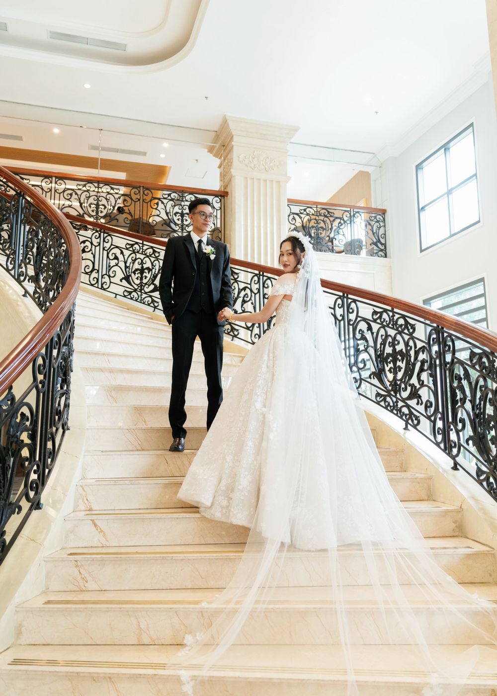 Elegant couple pose on grand marble staircase with ornate wrought iron railings during wedding photoshoot.