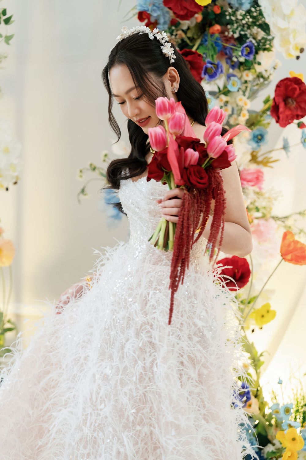 Bride in feathered white gown holds vibrant pink and red floral bouquet against backdrop of colorful flowers.