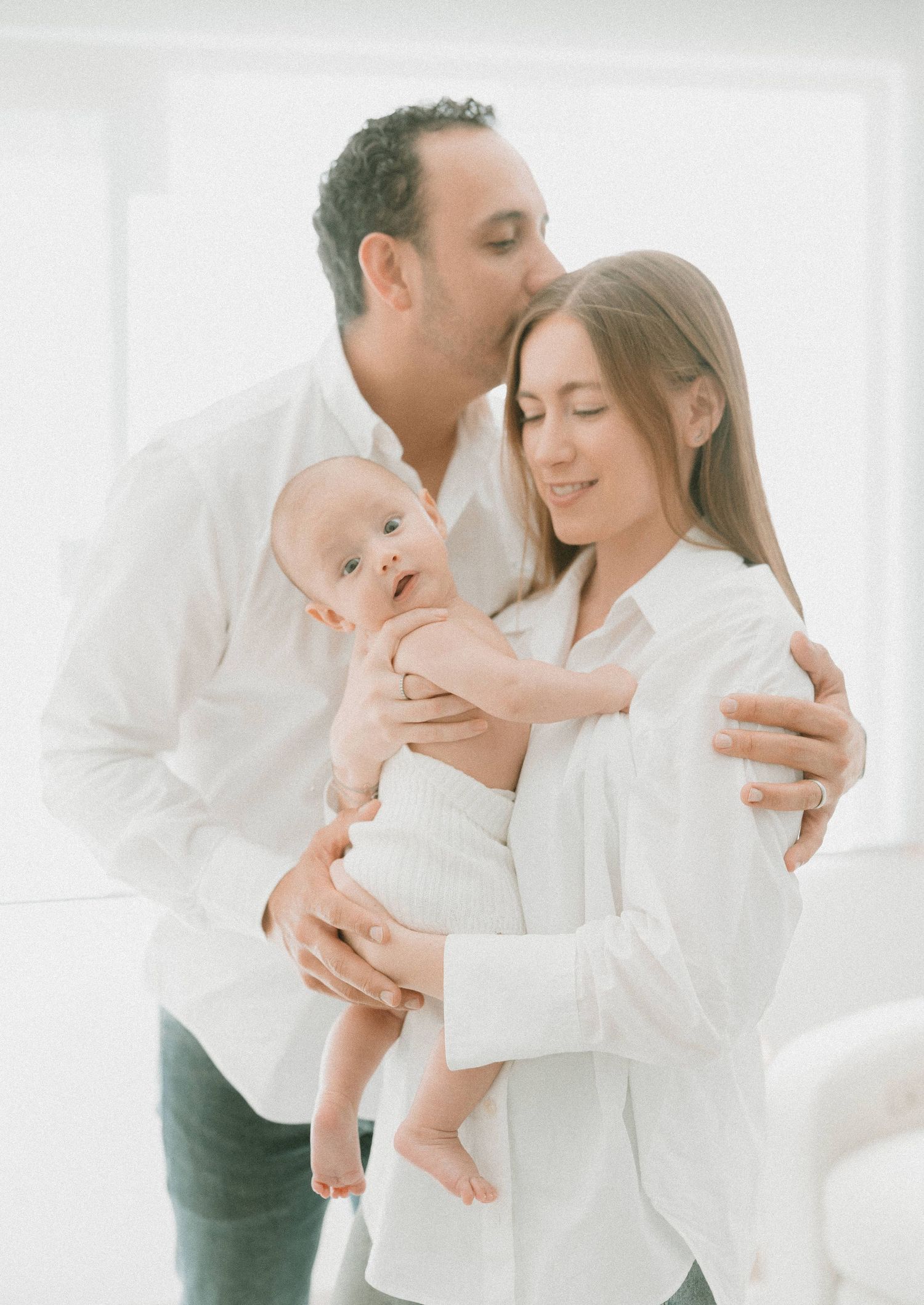Tender family moment with parents in white embracing their baby in bright minimalist setting.