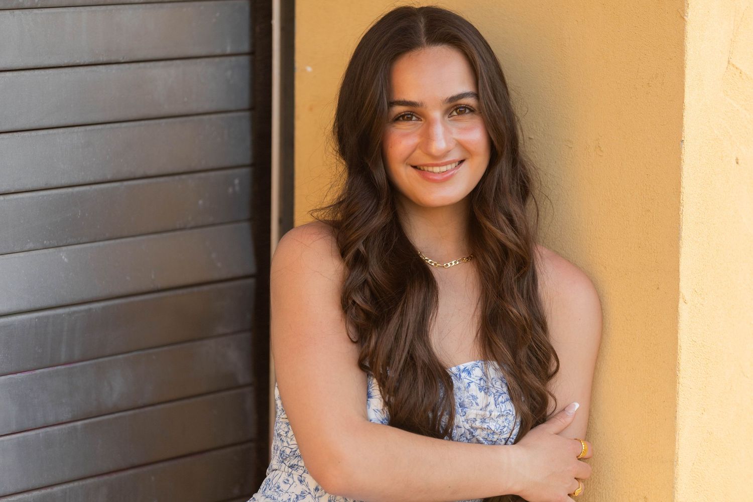 Portrait of a young woman with long dark hair smiling against a yellow wall with dark shutters.